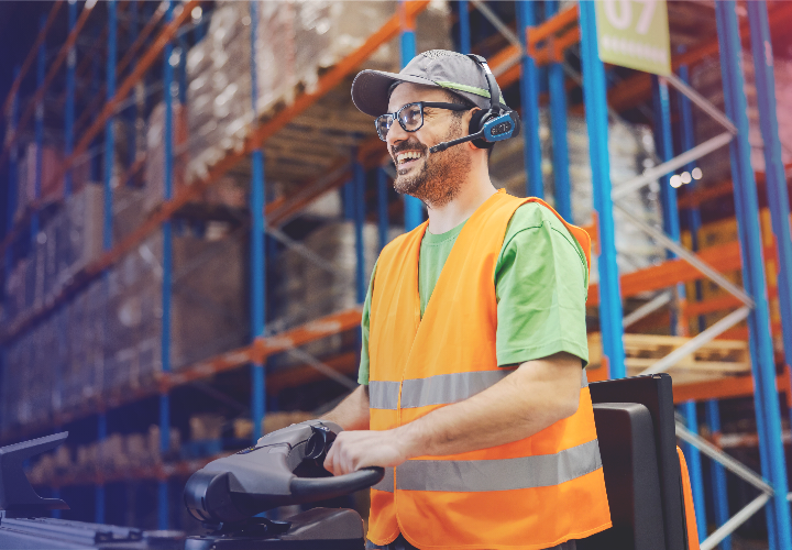 A warehouse worker drives a truck with a safety vest and headset on 