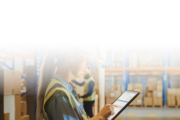 A female warehouse worker using a tablet to check inventory through warehouse management software, standing near organized shelves.