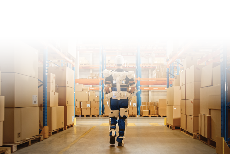 A warehouse worker wearing a robotic exosuit walks through a warehouse filled with shelves of organized inventory.