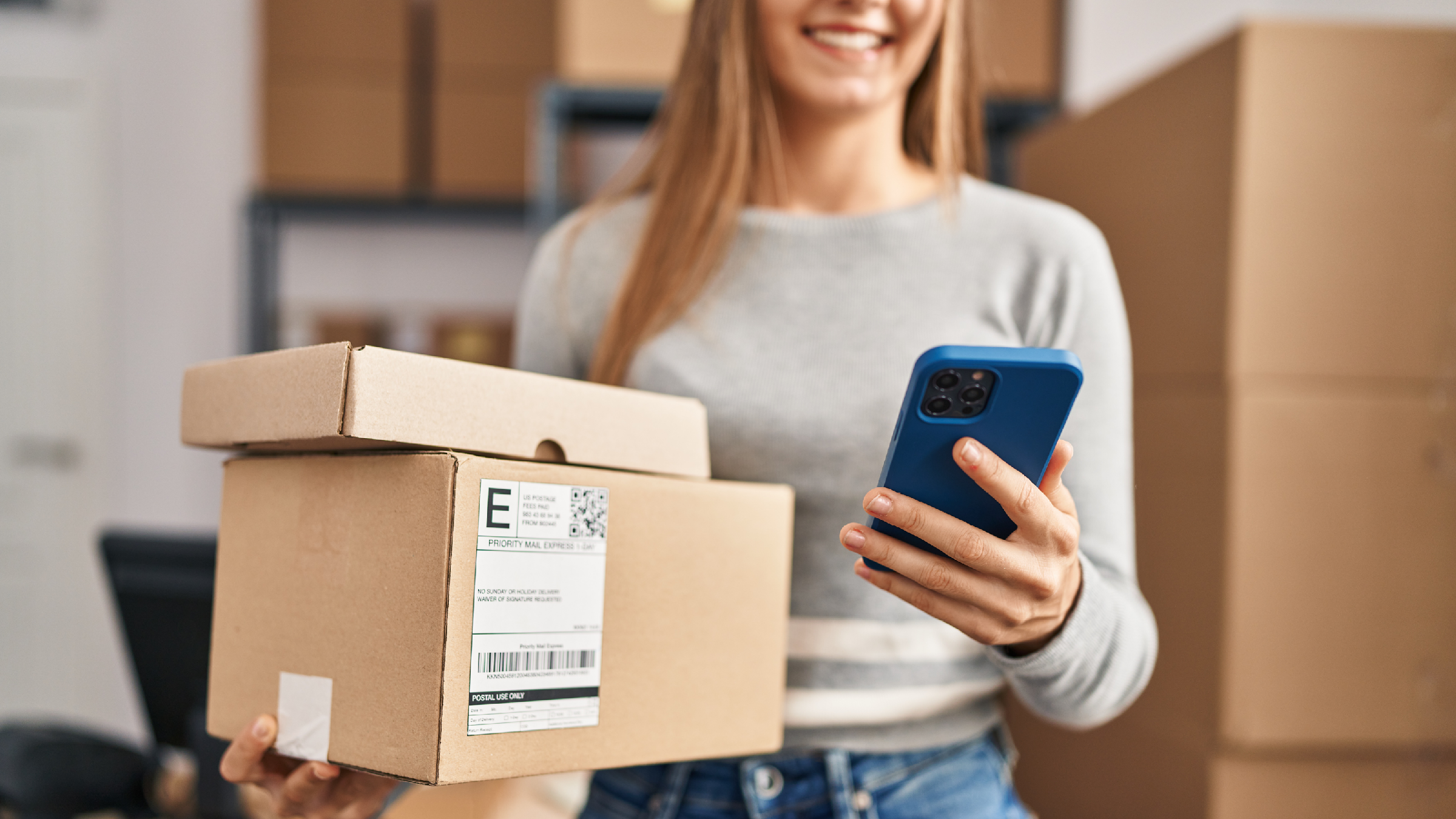 Happy woman balancing packages in one hand while holding a mobile phone in the other