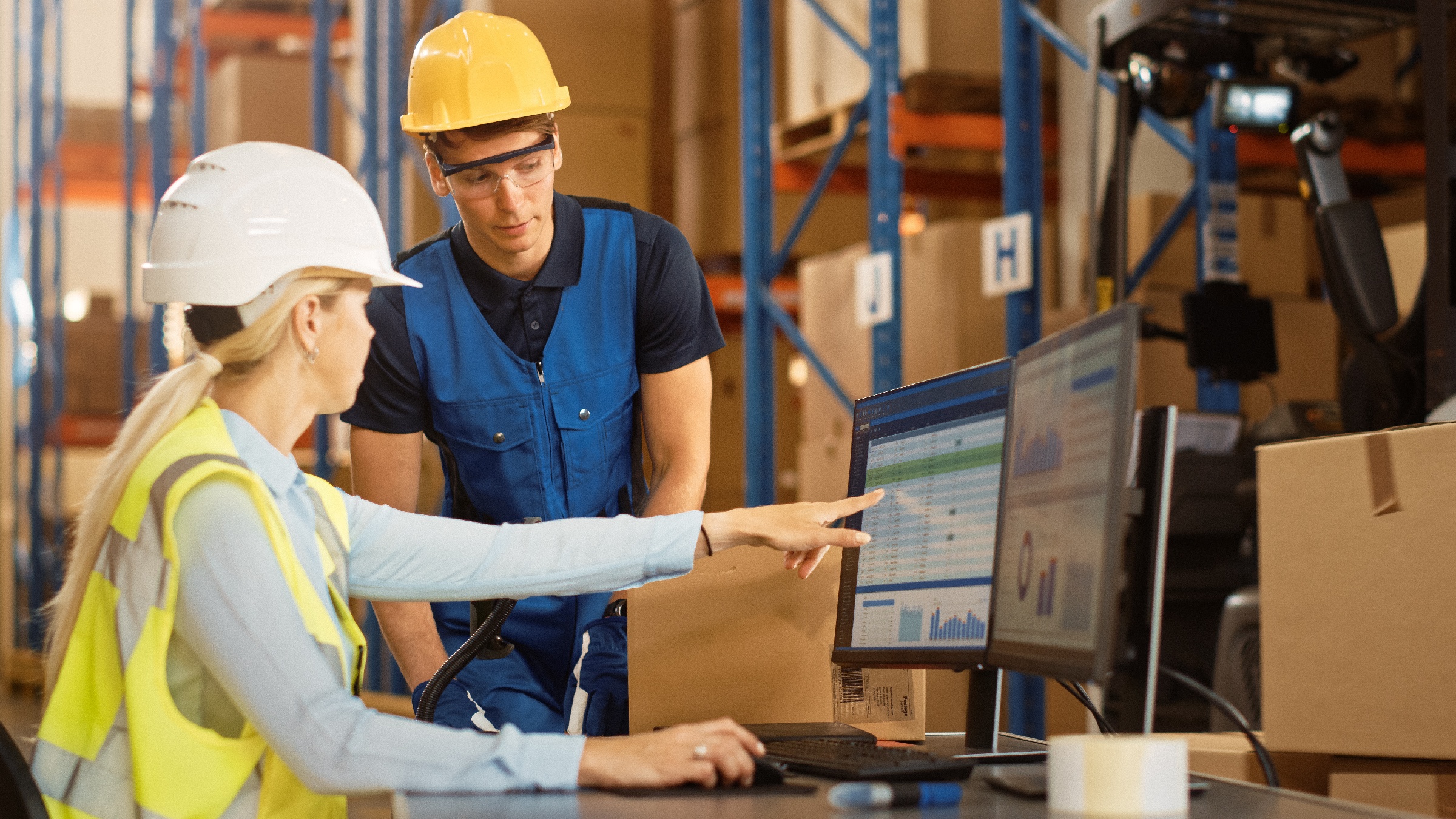 Two warehouse workers discuss operations, viewing ERP and WMS software on a screen in a warehouse.