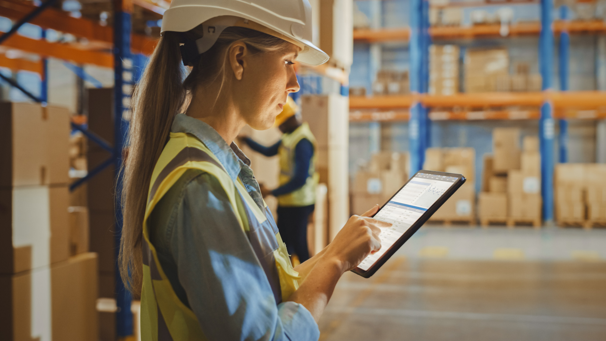 A female warehouse worker stands in the warehouse wearing a protective helmet and uses a tablet with the best warehouse management system