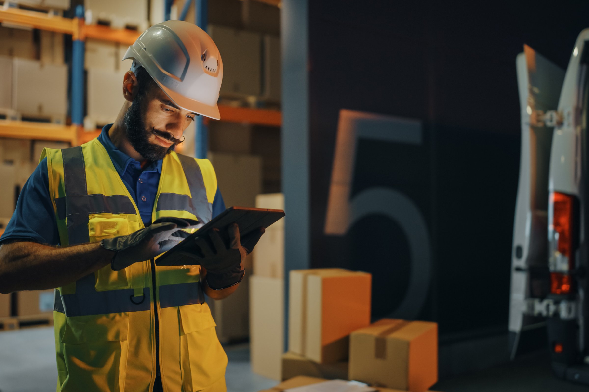 Man with safety vest and helmet looking at e tablet standing in a warehouse